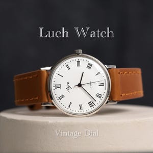 Puede incluir: Un reloj Luch con una esfera vintage blanca y marcadores de hora en números romanos. El reloj tiene una correa de cuero marrón y está sobre una superficie blanca.