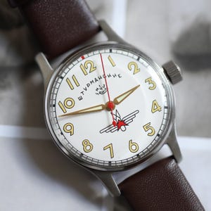 May include: A vintage wristwatch with a white face, featuring gold numerals and hands. The watch has a red second hand and a stainless steel case, paired with a brown leather strap. The watch face includes Cyrillic text and a red star emblem.
