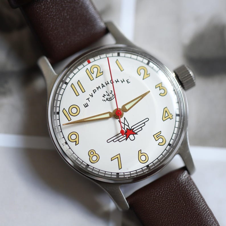 May include: A vintage wristwatch with a white face, featuring gold numerals and hands. The watch has a red second hand and a stainless steel case, paired with a brown leather strap. The watch face includes Cyrillic text and a red star emblem.