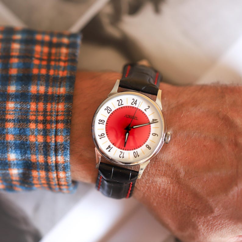 May include: A silver wristwatch with a red and white face and black leather band. The watch face displays a 24-hour clock with white numerals and a red center. The band has red stitching.