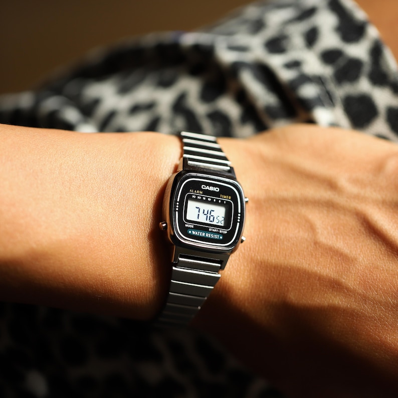 K&ouml;nnte beinhalten: Eine silberfarbene Digitalarmbanduhr mit rechteckigem Zifferblatt und Metallgliederarmband. Das schwarze Zifferblatt zeigt den Markennamen CASIO, die W&ouml;rter ALARM und TIMER sowie den Text WATER RESIST. Das Digitaldisplay zeigt die Uhrzeit 7:46.