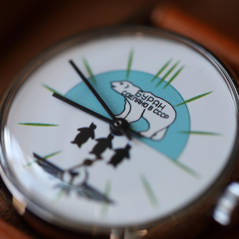 Puede incluir: Un reloj de pulsera con esfera blanca que presenta un oso polar en un c&iacute;rculo azul claro, con el texto "Бурaн Сделано en la URSS". Debajo del c&iacute;rculo hay tres siluetas negras. El reloj tiene agujas negras y una caja plateada.