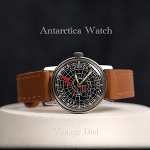 May include: A vintage wristwatch with a black dial featuring a map of Antarctica and the text "АRКТИКА" and "СЕВЕРНЫЙ ПОЛЮС". The watch has a silver case and a brown leather band.