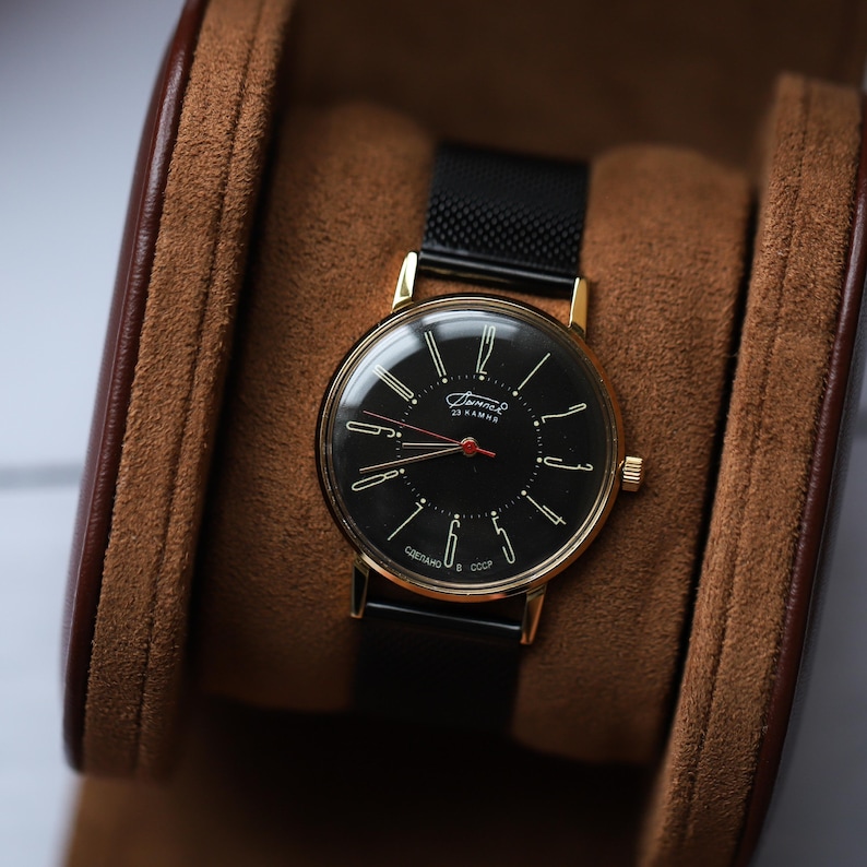 May include: A vintage wristwatch with a black face, gold-tone case, and black mesh band. The watch face features gold hands and markers, with the text "Зимний" and "23 KAMHE". The watch is displayed in a brown watch box.