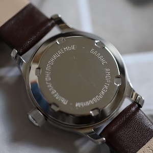 May include: Close-up of a vintage wristwatch with a silver-colored case and a brown leather strap. The watch back has engraved Cyrillic text. The watch is a classic timepiece.