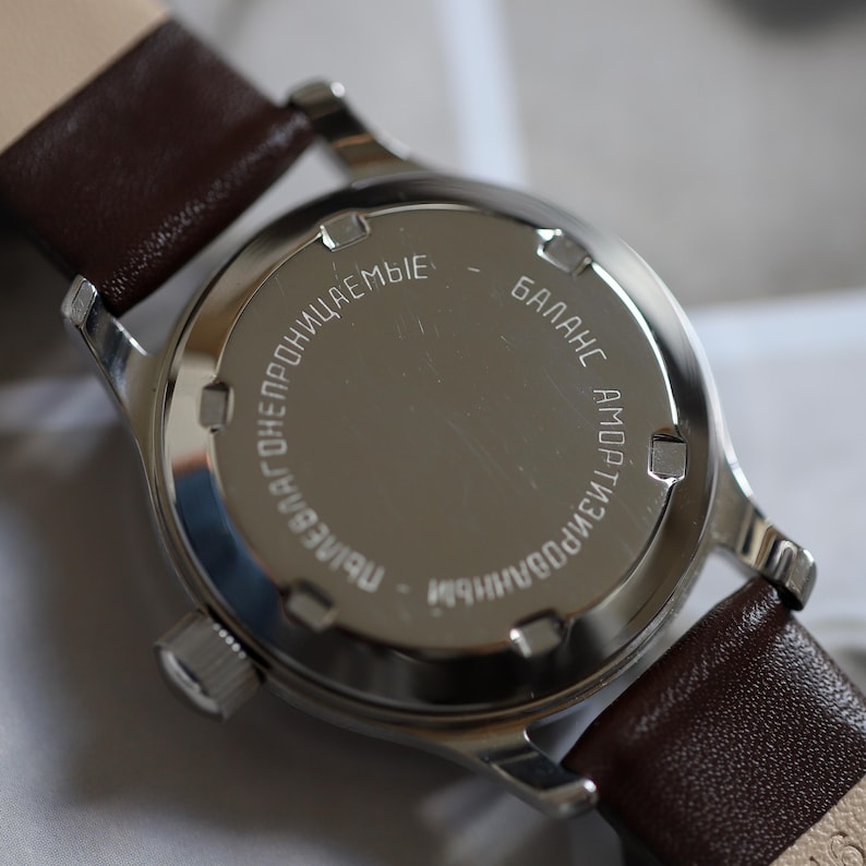 May include: Close-up of a vintage wristwatch with a silver-colored case and a brown leather strap. The watch back has engraved Cyrillic text. The watch is a classic timepiece.