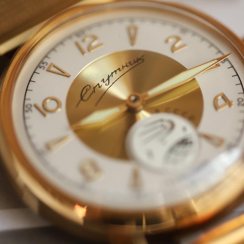 K&ouml;nnte beinhalten: Eine goldfarbene Armbanduhr mit wei&szlig;em Zifferblatt, goldenen Ziffern und einer zentralen goldenen Scheibe. Das Zifferblatt zeigt den kyrillischen Text Спутник und ein kleines wei&szlig;es Hilfszifferblatt mit einer Satellitengrafik. Goldene Zeiger.