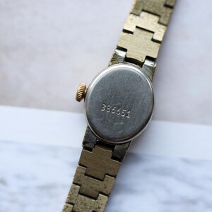 Puede incluir: Primer plano de un reloj de pulsera vintage en tono dorado. El reloj tiene una esfera redonda plateada con el n&uacute;mero "386651" grabado en la parte posterior. La correa del reloj es de dise&ntilde;o segmentado en tono dorado. La corona del reloj es dorada.