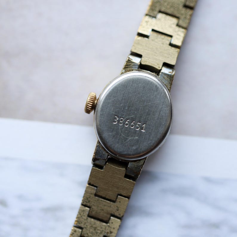 Puede incluir: Primer plano de un reloj de pulsera vintage en tono dorado. El reloj tiene una esfera redonda plateada con el n&uacute;mero "386651" grabado en la parte posterior. La correa del reloj es de dise&ntilde;o segmentado en tono dorado. La corona del reloj es dorada.