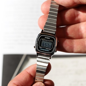 K&ouml;nnte beinhalten: Eine silberfarbene Digitalarmbanduhr mit rechteckigem Zifferblatt und Metallgliederarmband. Das schwarze Zifferblatt zeigt den Markennamen CASIO, die Begriffe ALARM, TIMER und WATER RESIST sowie eine digitale Zeitanzeige von 6:57 und 20 Sekunden.