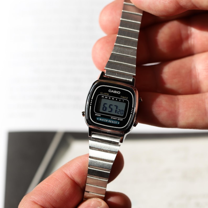 K&ouml;nnte beinhalten: Eine silberfarbene Digitalarmbanduhr mit rechteckigem Zifferblatt und Metallgliederarmband. Das schwarze Zifferblatt zeigt den Markennamen CASIO, die Begriffe ALARM, TIMER und WATER RESIST sowie eine digitale Zeitanzeige von 6:57 und 20 Sekunden.