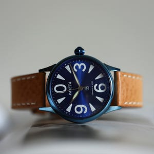 May include: A blue wristwatch with a tan leather strap. The watch face is dark blue with white numerals and hands. The word "PAKETA" is printed on the watch face.