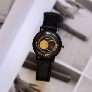 Puede incluir: Un reloj de pulsera negro con una esfera redonda que presenta un dise&ntilde;o de c&iacute;rculo y anillo de color dorado. El reloj tiene una correa de cuero negro y la palabra "PAKETA" est&aacute; impresa en la esfera. El reloj est&aacute; sobre un fondo borroso.