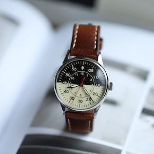 May include: A vintage-style wristwatch with a black and cream-colored face and a brown leather strap. The watch face features black numerals and a red second hand. The watch case is silver-toned. The strap is stitched with cream-colored thread.