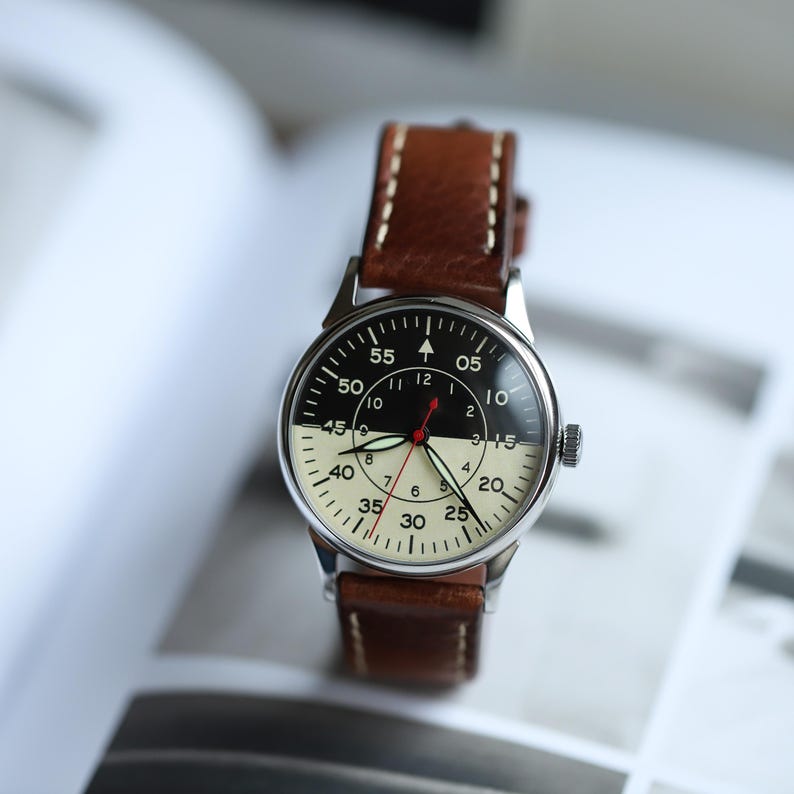 May include: A vintage-style wristwatch with a black and cream-colored face and a brown leather strap. The watch face features black numerals and a red second hand. The watch case is silver-toned. The strap is stitched with cream-colored thread.