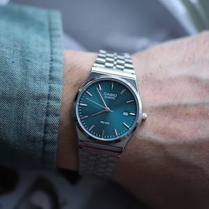 May include: A silver Casio Quartz watch with a teal face and date display. The watch has a stainless steel band and is on a wrist. The watch face has the text "CASIO QUARTZ" and "WR 50M".