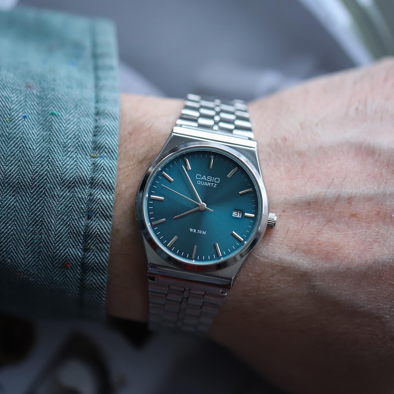 May include: A silver Casio Quartz watch with a teal face and date display. The watch has a stainless steel band and is on a wrist. The watch face has the text "CASIO QUARTZ" and "WR 50M".