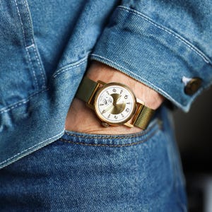 K&ouml;nnte beinhalten: Eine goldfarbene Armbanduhr mit rundem wei&szlig;em Zifferblatt, goldfarbenen Zeigern und arabischen Ziffern. Die Uhr verf&uuml;gt &uuml;ber ein Metall-Mesh-Armband und ein kreisf&ouml;rmiges Hilfszifferblatt. Sie wird am Handgelenk unter einer blauen Jeansjacke getragen.