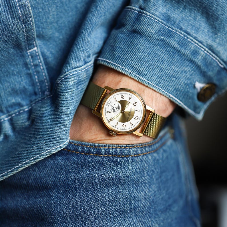 K&ouml;nnte beinhalten: Eine goldfarbene Armbanduhr mit rundem wei&szlig;em Zifferblatt, goldfarbenen Zeigern und arabischen Ziffern. Die Uhr verf&uuml;gt &uuml;ber ein Metall-Mesh-Armband und ein kreisf&ouml;rmiges Hilfszifferblatt. Sie wird am Handgelenk unter einer blauen Jeansjacke getragen.