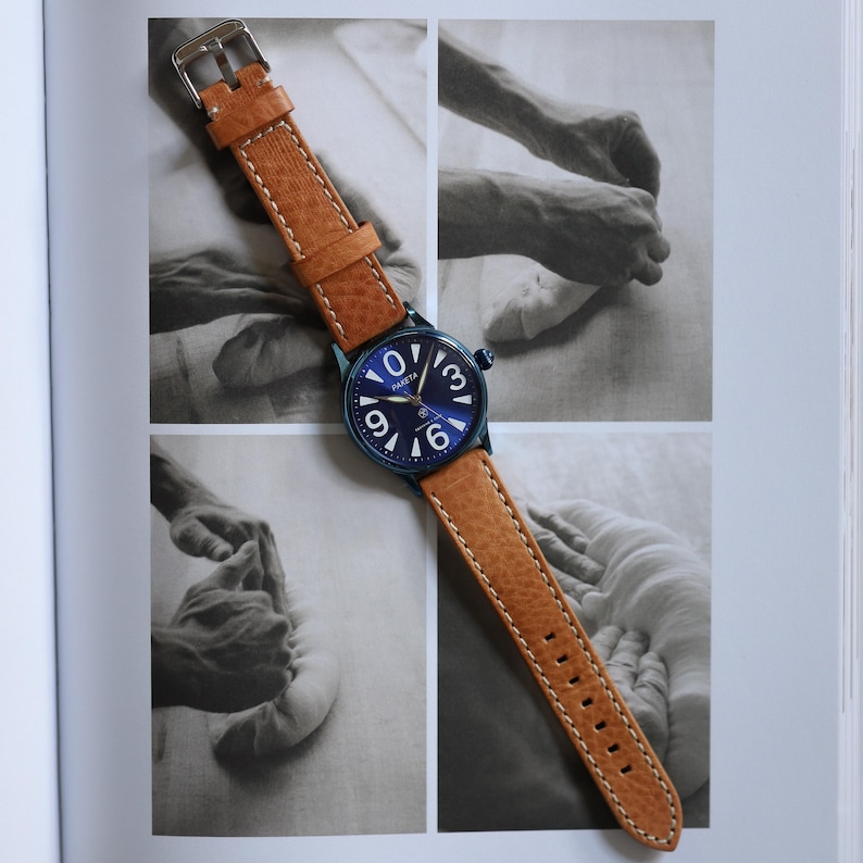 May include: A wristwatch featuring a brown leather strap and a blue face with white numerals and hands. The strap is stitched with white thread and has a silver buckle. The watch is displayed on a book with black and white images.