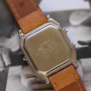 May include: Close-up of a Casio AQ-230 watch with a stainless steel back and a brown leather strap. The watch face is not visible. The text on the back reads "CASIO", "STAINLESS STEEL BACK", and "WATER RESISTANT".