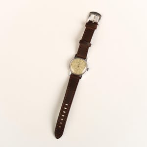 Pode incluir: Uma pulseira de rel&oacute;gio de couro marrom com um mostrador prateado. O mostrador do rel&oacute;gio tem um fundo branco com n&uacute;meros e ponteiros pretos. A pulseira do rel&oacute;gio tem detalhes de costura.