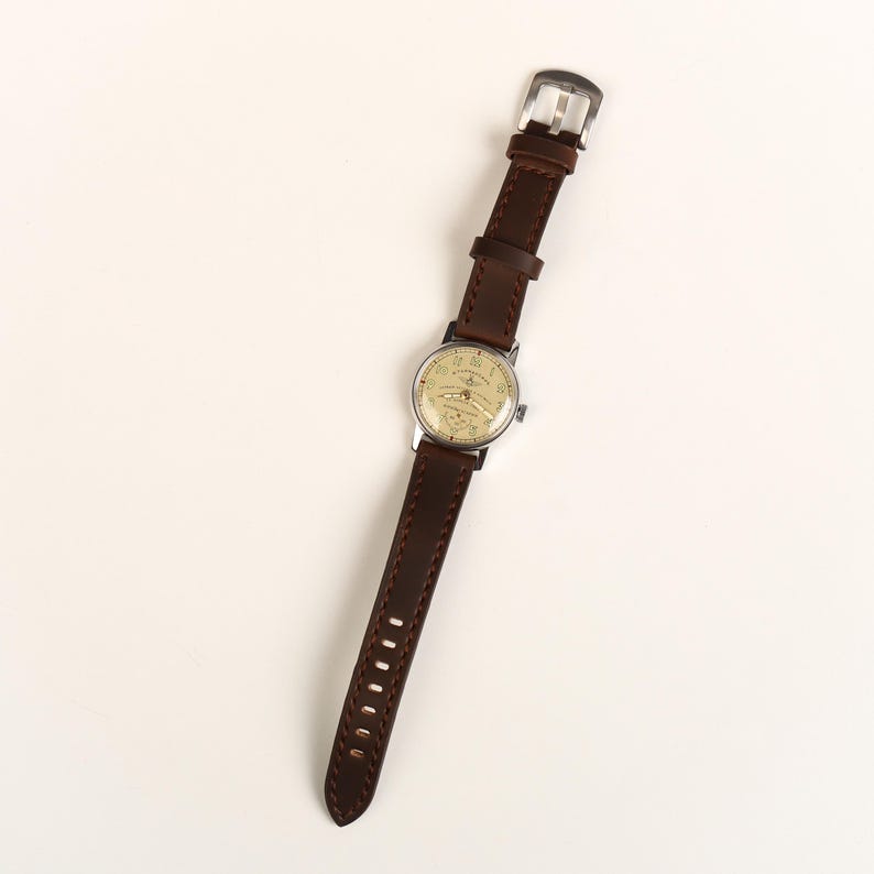 Pode incluir: Uma pulseira de rel&oacute;gio de couro marrom com um mostrador prateado. O mostrador do rel&oacute;gio tem um fundo branco com n&uacute;meros e ponteiros pretos. A pulseira do rel&oacute;gio tem detalhes de costura.
