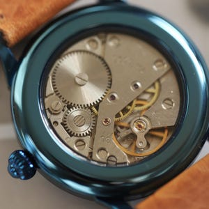 May include: Close-up of a teal wristwatch revealing its mechanical workings. The watch has a brown leather strap and a teal bezel. The internal gears and components are visible, showcasing the watch's intricate design and craftsmanship.