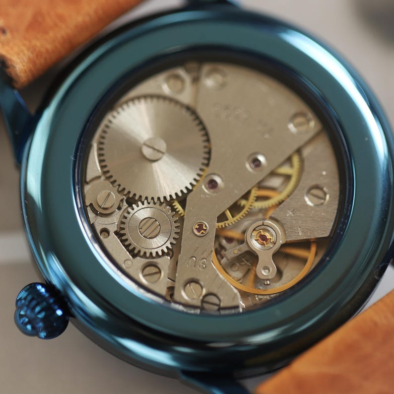 May include: Close-up of a teal wristwatch revealing its mechanical workings. The watch has a brown leather strap and a teal bezel. The internal gears and components are visible, showcasing the watch's intricate design and craftsmanship.