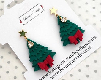 May include: A pair of green Christmas tree earrings with red bows and gold stars. The earrings are on a white card with the text "Boutique Crafts" and website information.