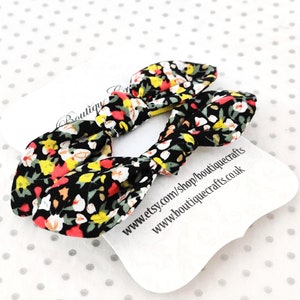 May include: Two fabric hair bows with a floral pattern in black, red, yellow, and white. The bows are tied in a knot shape. A white tag with text is visible in the background.