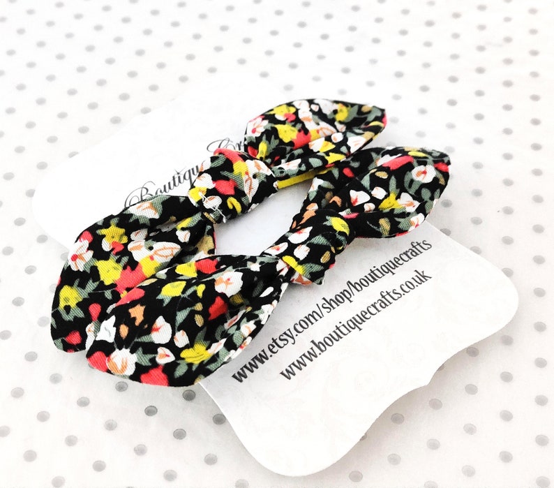 May include: Two fabric hair bows with a floral pattern in black, red, yellow, and white. The bows are tied in a knot shape. A white tag with text is visible in the background.