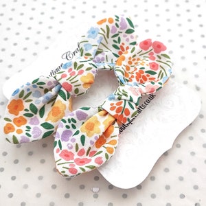 May include: Two floral print hair bows with a white background. The bows are made of fabric with a pattern of orange, pink, blue, and purple flowers on a white background. The bows are on white tags.