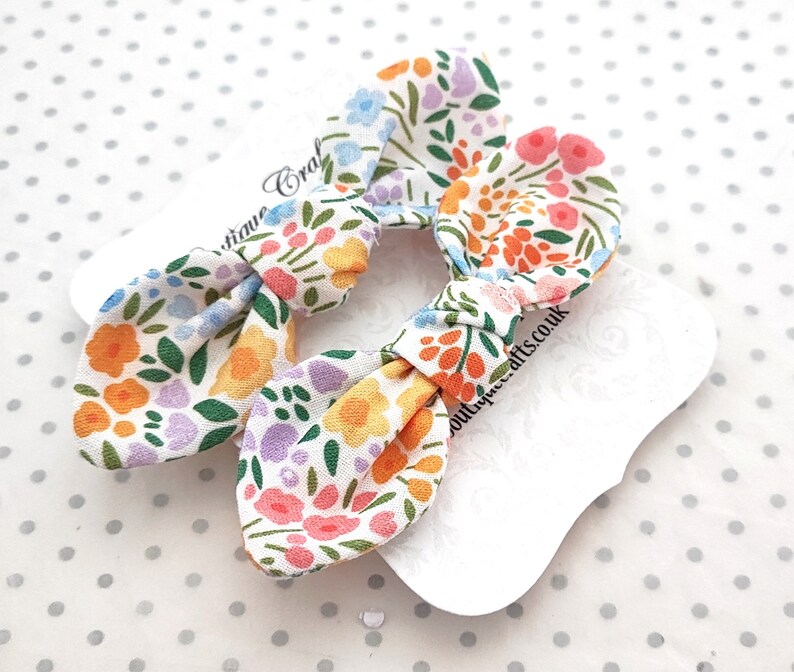 May include: Two floral print hair bows with a white background. The bows are made of fabric with a pattern of orange, pink, blue, and purple flowers on a white background. The bows are on white tags.