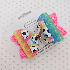 May include: A vibrant hair bow with a soccer ball design. The bow is made up of layers of glittery pink, yellow, and blue, and has a gold-coloured clip. It's attached to a white tag with the text "Boutique Crafts".