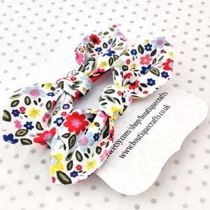 May include: Two floral print hair bows with a white background and colorful flowers in red, blue, pink, and yellow. The bows are tied in a knot. A white tag with text is visible.