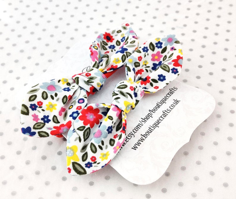 May include: Two floral print hair bows with a white background and colorful flowers in red, blue, pink, and yellow. The bows are tied in a knot. A white tag with text is visible.