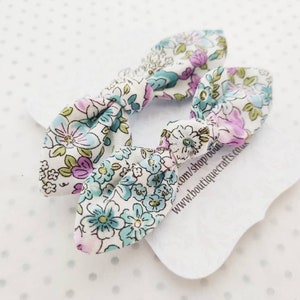 May include: Two floral patterned hair bows. The bows are white with a pattern of blue, purple, and green flowers. The bows are tied in a knot shape. The bows are on a white tag with text that reads www.boutiquecrafts.co.uk.