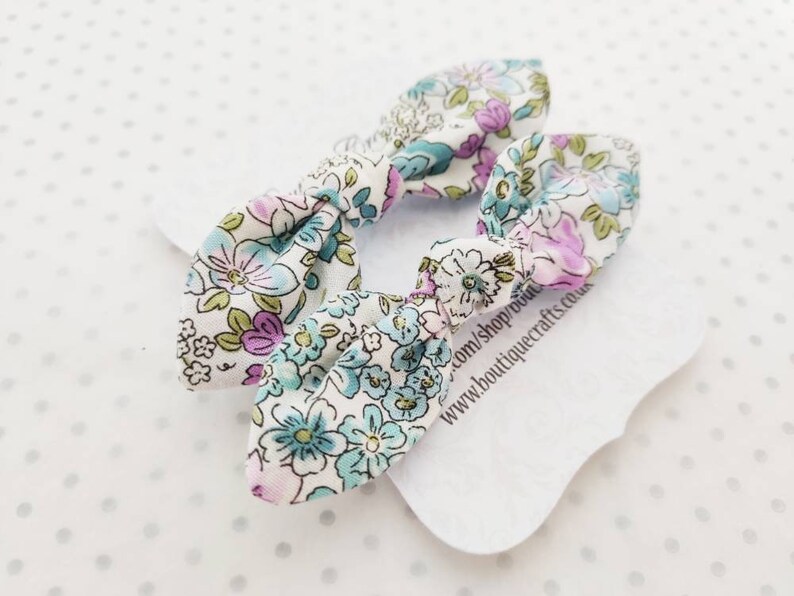 May include: Two floral patterned hair bows. The bows are white with a pattern of blue, purple, and green flowers. The bows are tied in a knot shape. The bows are on a white tag with text that reads www.boutiquecrafts.co.uk.