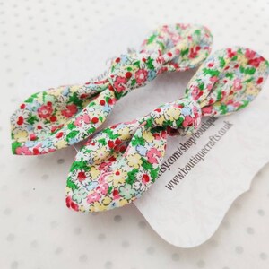 May include: Two floral fabric hair bows with a colorful pattern of red, pink, yellow, and green flowers on a white background. The bows are tied in a knot shape. A white tag with text is visible.
