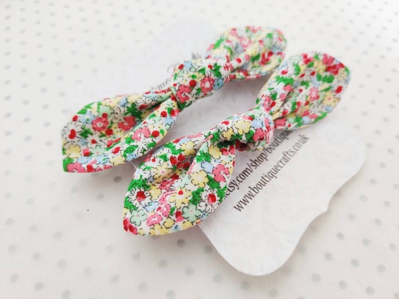 May include: Two floral fabric hair bows with a colorful pattern of red, pink, yellow, and green flowers on a white background. The bows are tied in a knot shape. A white tag with text is visible.