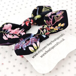 May include: Two black fabric hair bows with a floral pattern in pink, purple, and yellow. The bows are knotted in the center. A white tag with text is visible beneath the bows.