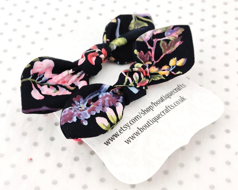 May include: Two black fabric hair bows with a floral pattern in pink, purple, and yellow. The bows are knotted in the center. A white tag with text is visible beneath the bows.