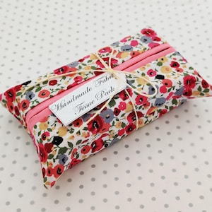 May include: A floral fabric tissue pack with a pink accent and a tag that reads "Handmade Fabric Tissue Pack". The fabric features red, yellow, and blue flowers on a white background. The tissue pack is tied with string.