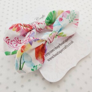 May include: Two floral print hair bows with a white background. The bows feature a colorful pattern of flowers and leaves in shades of pink, red, green, and blue. Each bow is tied in a knot. The bows are on a white tag with text.