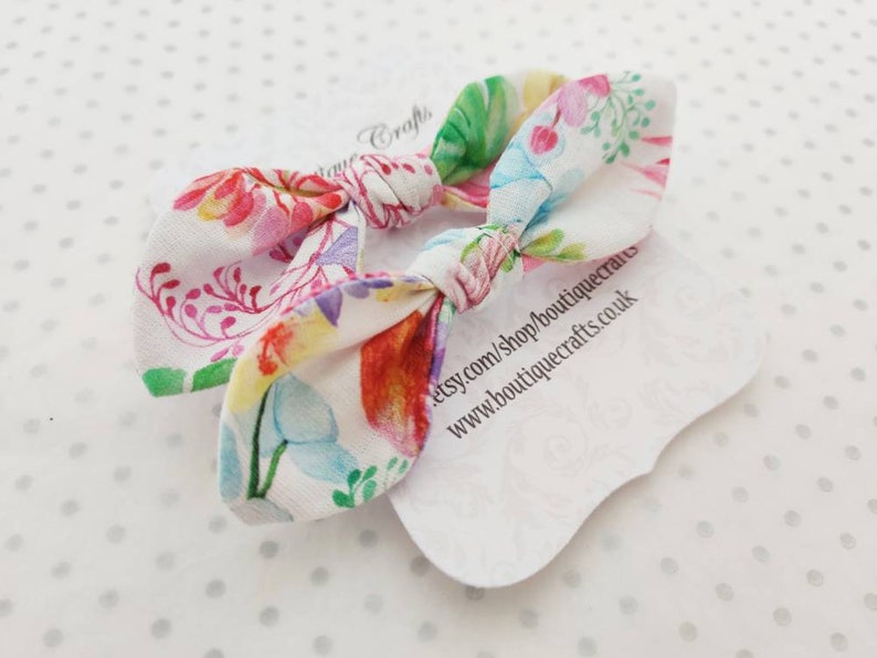 May include: Two floral print hair bows with a white background. The bows feature a colorful pattern of flowers and leaves in shades of pink, red, green, and blue. Each bow is tied in a knot. The bows are on a white tag with text.
