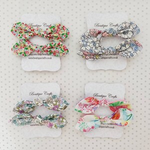 May include: Four pairs of floral fabric hair bows on white cards. Each bow features a different colorful floral pattern. The cards have the text "Boutique Crafts" and a website address.