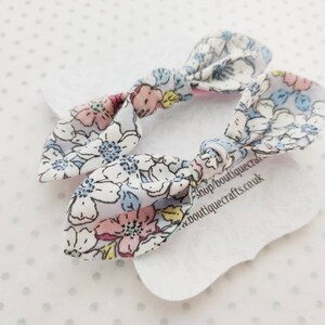 May include: Two floral patterned hair bows. The bows are white with a pattern of pink, blue, and yellow flowers. The bows are tied in a knot and are on a white tag with text.