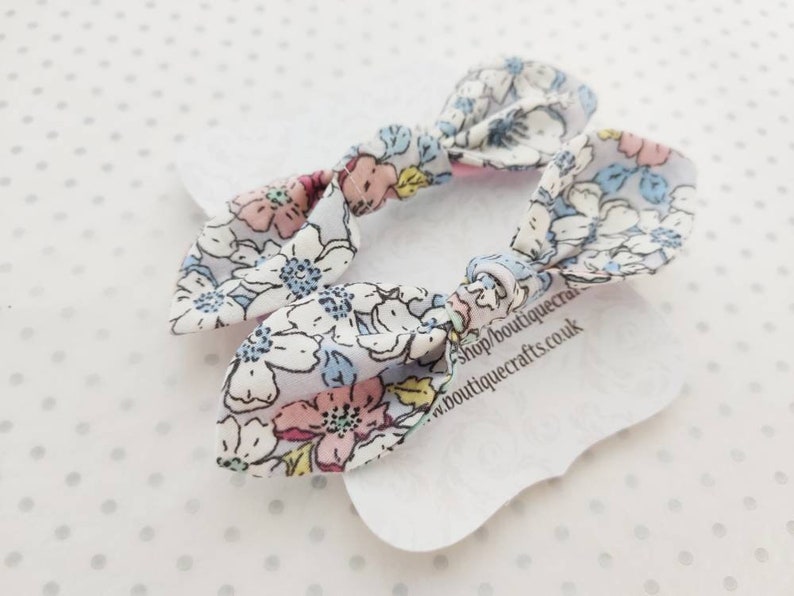 May include: Two floral patterned hair bows. The bows are white with a pattern of pink, blue, and yellow flowers. The bows are tied in a knot and are on a white tag with text.