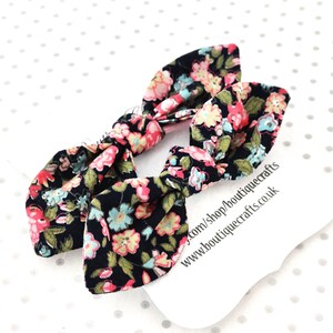 May include: Two black fabric hair bows with a floral pattern in pink, blue, and yellow. The bows are tied in a bunny ear style. A white tag with text is in the background.